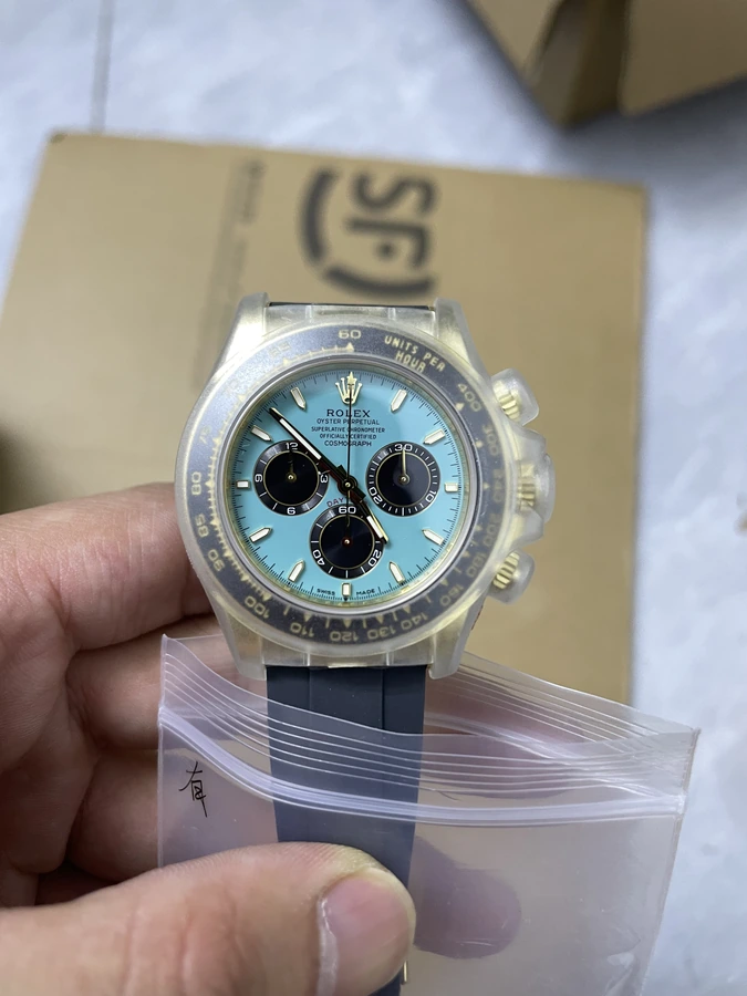 Light blue dial watch with three black subdials, a dark numbered bezel with gold markings and black strap, photographed in hand above clear packaging.