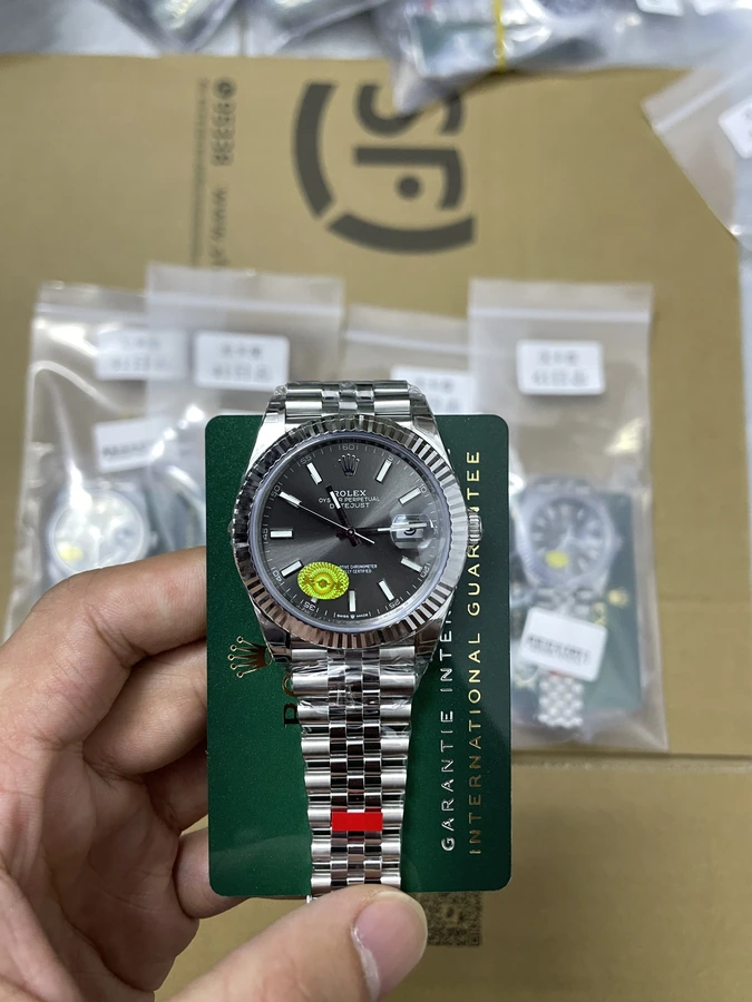 Gray dial watch with silver bracelet and fluted bezel, photographed in hand on a green presentation card.